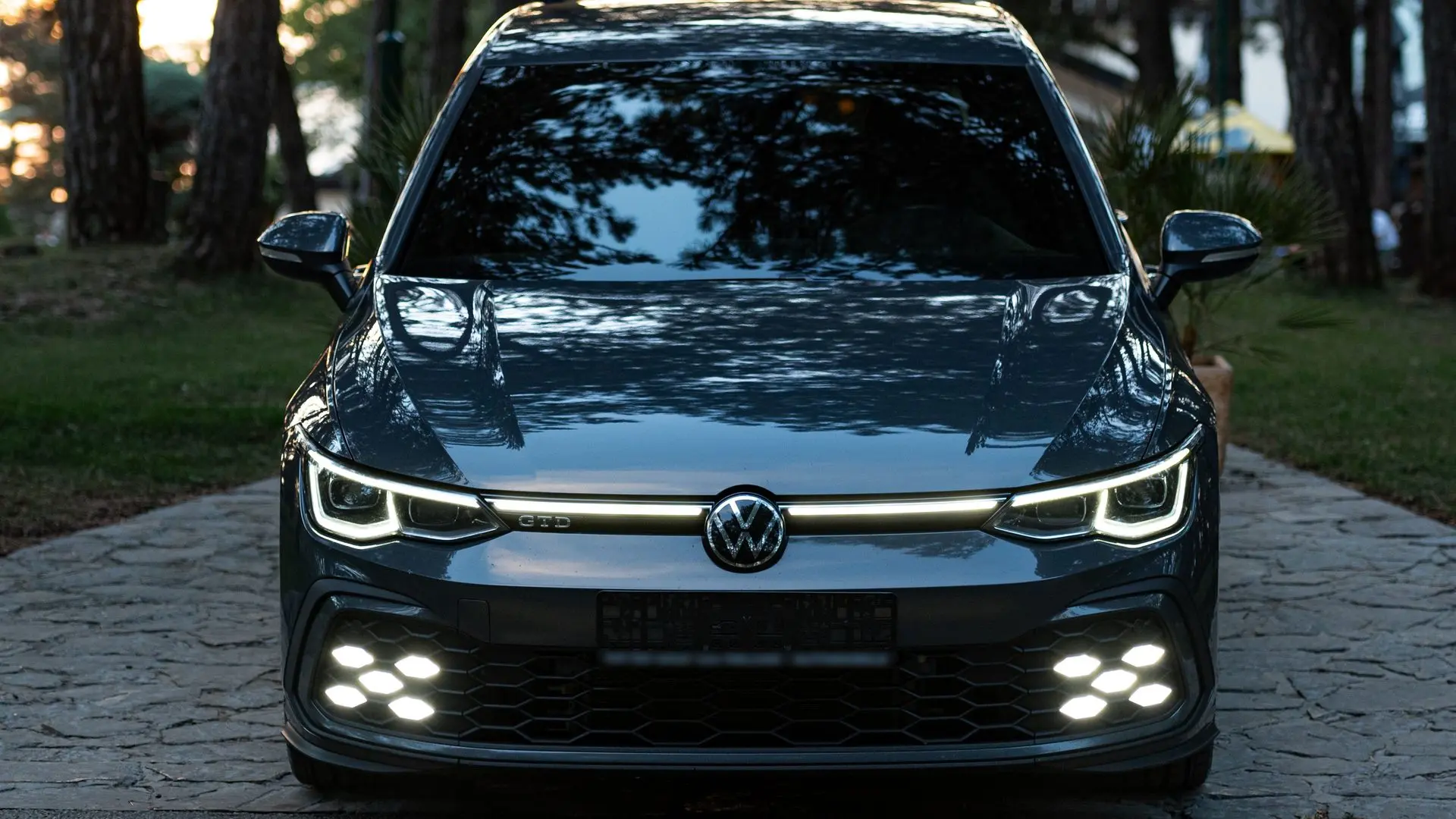 Alacakaranlıkta farları ve ızgara ışık bandı yanan koyu gri Volkswagen Golf GTD